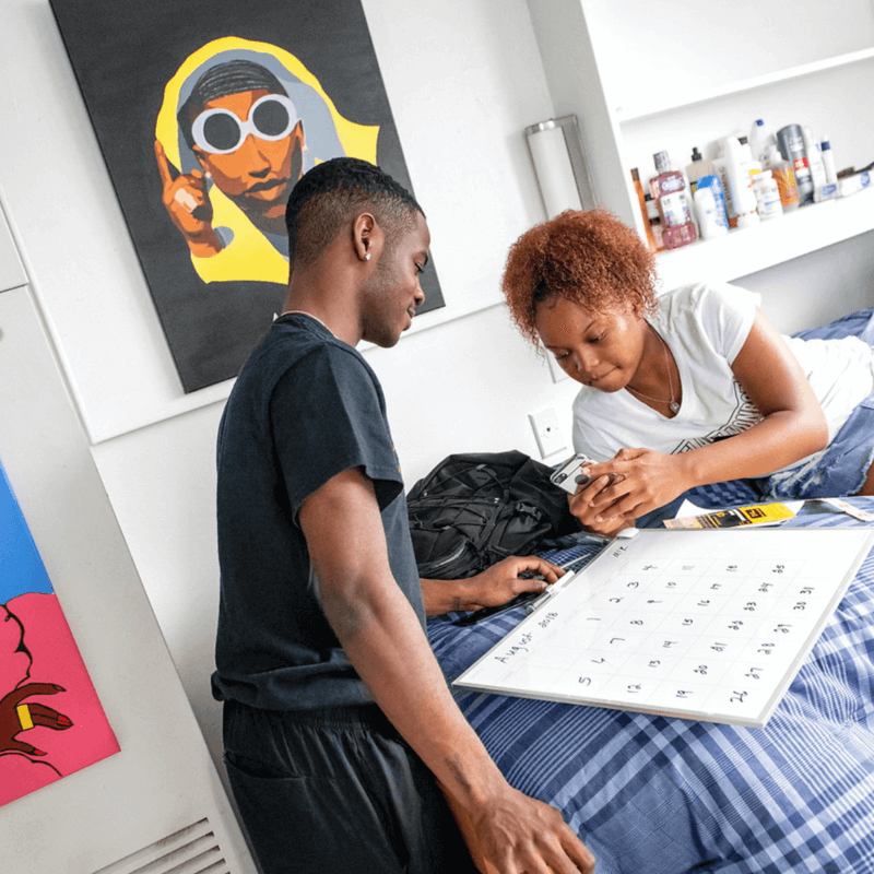 A howard student with a family member writing on a calendar in a dorm room.