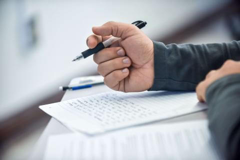 A close-up of a student's hand gripping a pencil over a piece of paper.