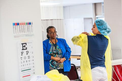 Two health professionals in a medical setting. One of them is putting on personal protective gear.